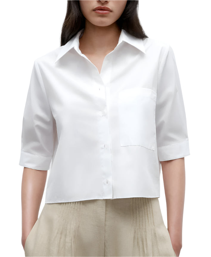 A woman wears the TWP Short Lived Top—a white, boxy fit, short-sleeved cotton poplin button-up shirt with a chest pocket—paired with beige pleated pants, shown from shoulders to hips.