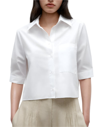 A woman wears the TWP Short Lived Top—a white, boxy fit, short-sleeved cotton poplin button-up shirt with a chest pocket—paired with beige pleated pants, shown from shoulders to hips.