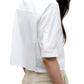 A woman wearing the TWP Short Lived Top—a white, boxy fit, short-sleeve collared shirt—paired with beige pants, is shown from the back and side against a white background.