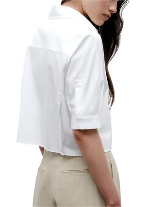 A woman wearing the TWP Short Lived Top—a white, boxy fit, short-sleeve collared shirt—paired with beige pants, is shown from the back and side against a white background.
