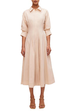 A woman in a Simkhai Signature Jazz dress in beige midi-length with short sleeves, a collar, and a pintucked waist, standing against a light gray background. She wears beige strappy sandals.