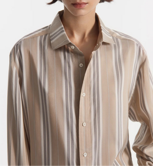 A person shown from shoulders to mid-torso wears the CO Standing Collar Silk Shirt, an oversized, beige-and-white striped button-down with the top button undone and elegant horn button detailing.