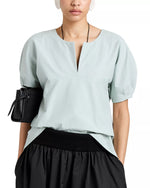 Someone wearing a light blue Proenza Schouler White Label Tia Top with a front slit neckline, paired with a black skirt and carrying a small black bag under their arm.