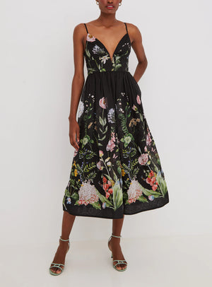 A person wearing the Cara Cara Pero Midi Dress, a black sleeveless fit-and-flare midi with floral prints, and green strappy heels stands against a plain white background.