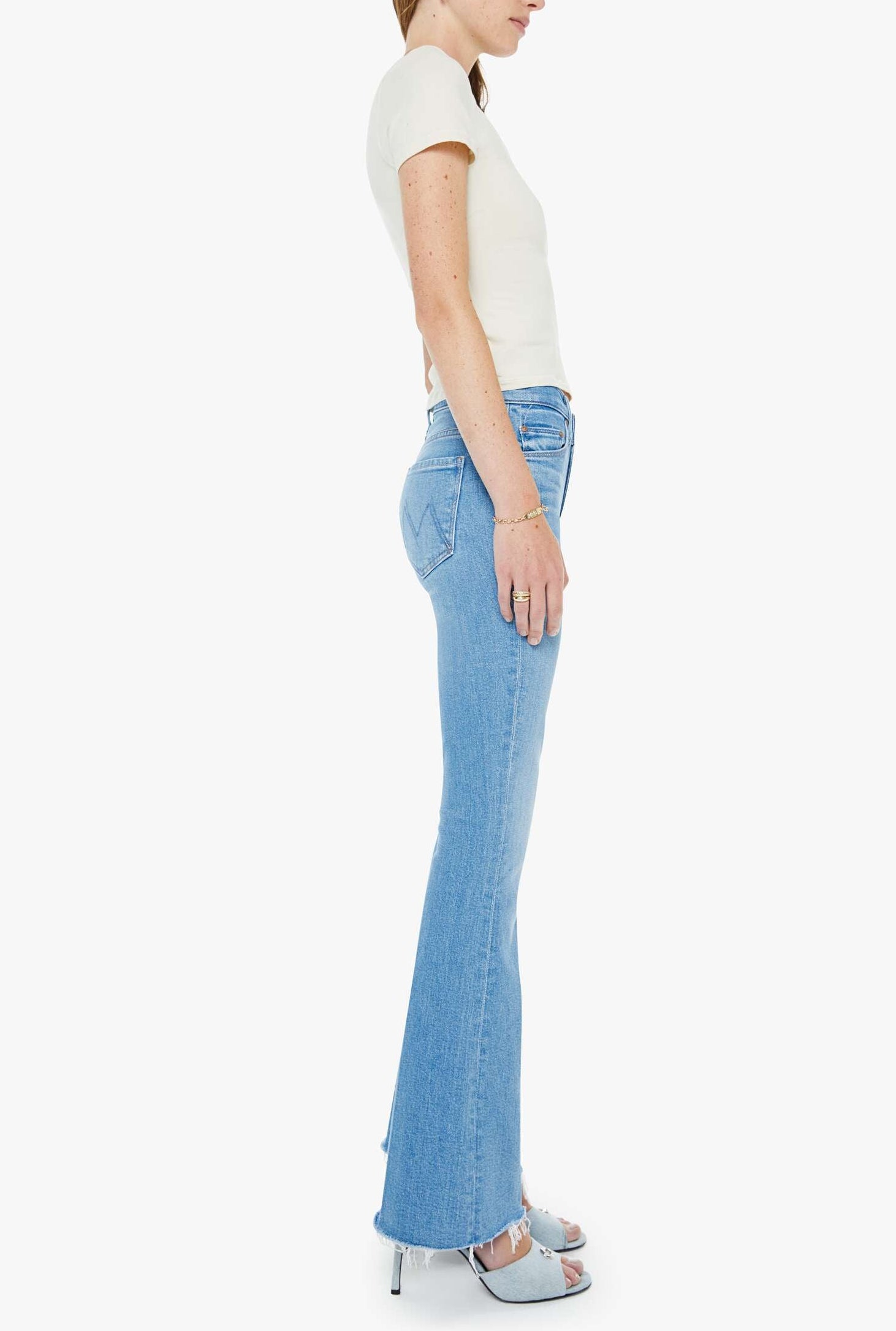 Woman in a short-sleeved white top, Mother’s The Weekender Fray mid-rise flare jeans crafted from recycled stretch denim, and heeled sandals stands in profile against a plain white background.