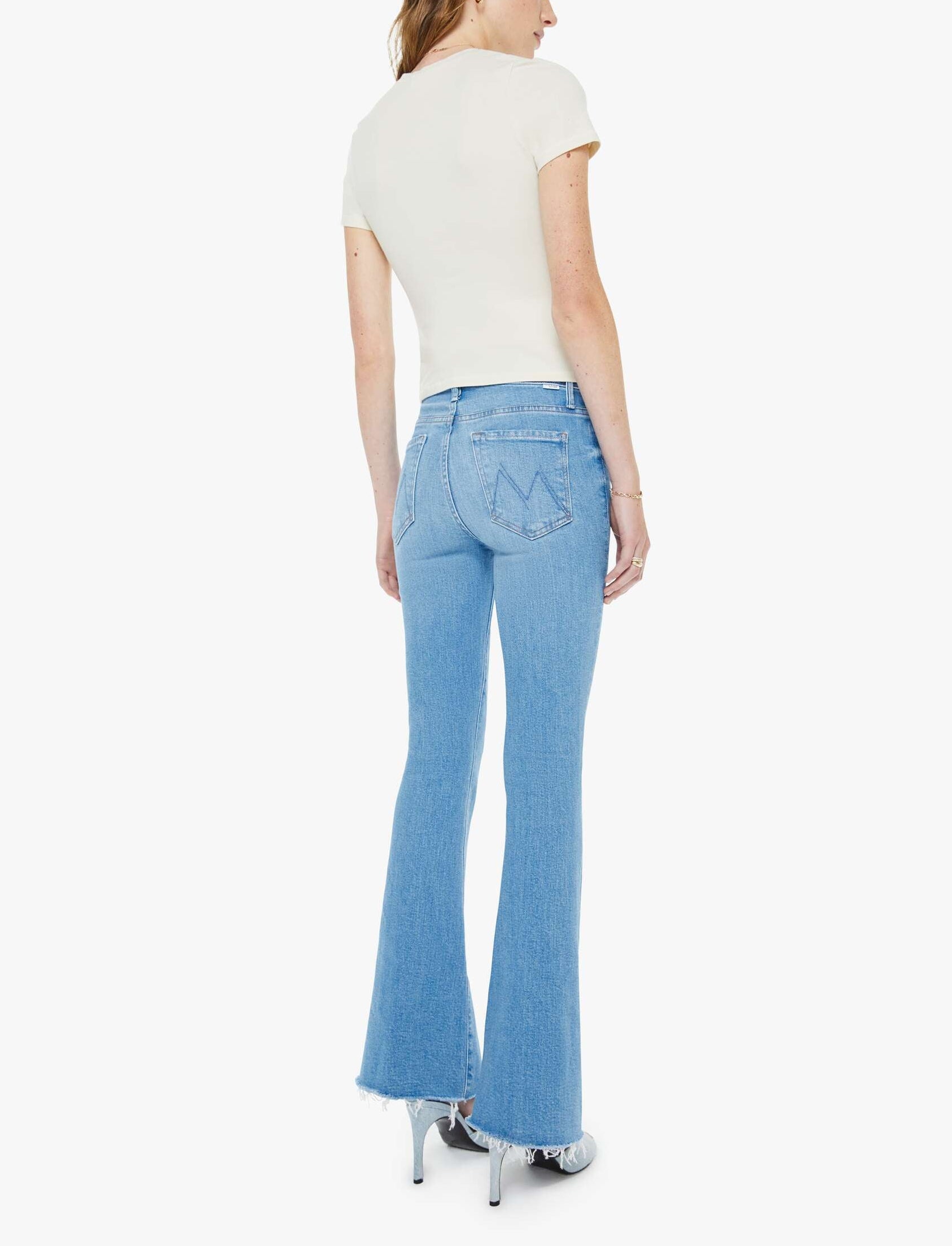 A person wearing a white short-sleeve top, The Weekender Fray light blue mid-rise flare jeans from Mother crafted from recycled stretch denim with raw hems, and silver high heels, stands with their back to the camera against a plain white background.