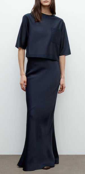 A woman in a fitted, floor-length My Endless Love Skirt by TWP and a loose black short-sleeve top with a chest pocket stands against a plain background.