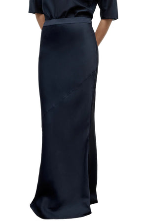 A person wearing the My Endless Love Skirt by TWP, a high-waisted, floor-length black satin maxi with diagonal seam detail, made from coated viscose linen, stands with hands behind their back.