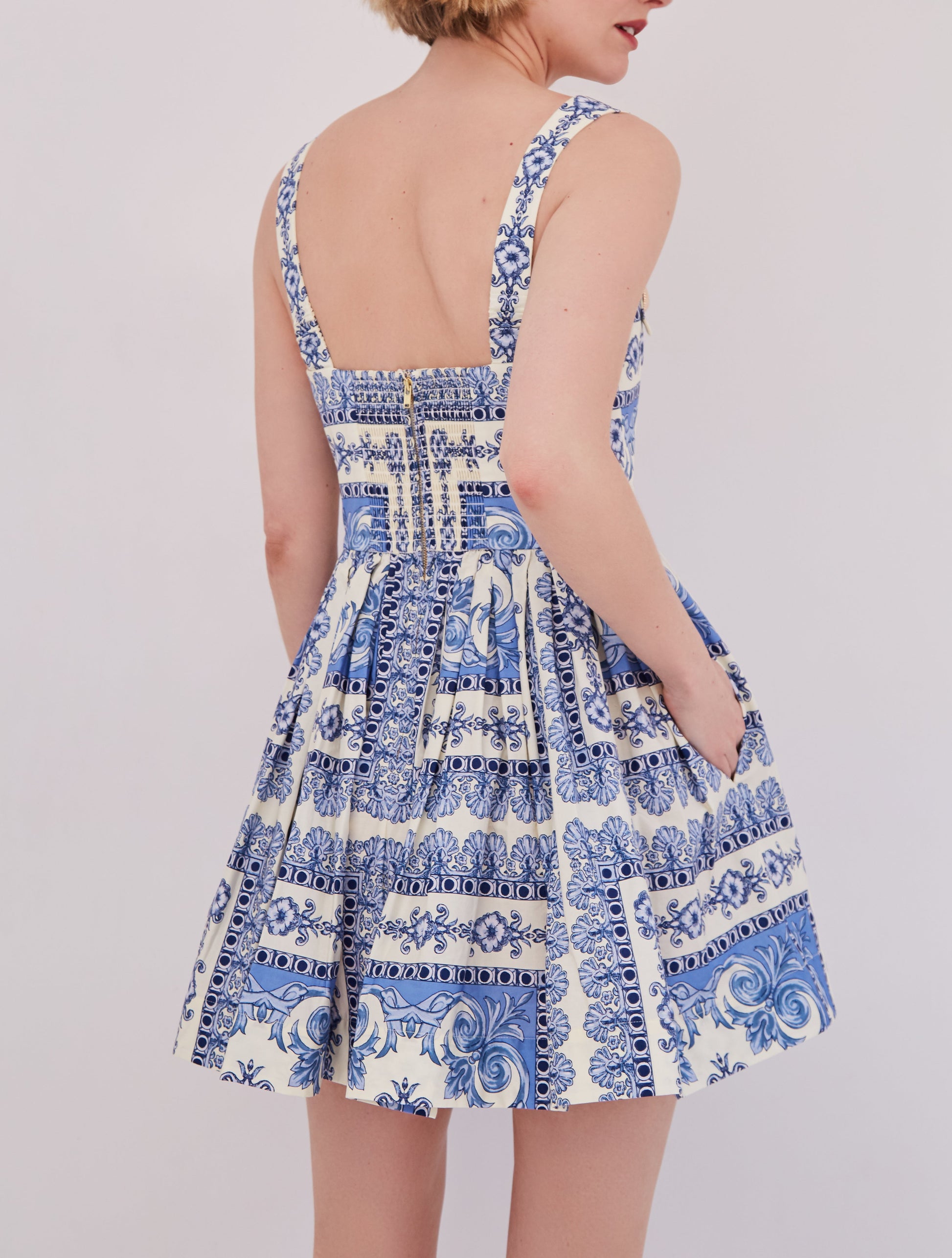 A woman stands facing away, wearing the Cara Cara Micaela Mini Dress—a blue and white patterned fit-and-flare style with a structured bodice, square back, and visible zipper.