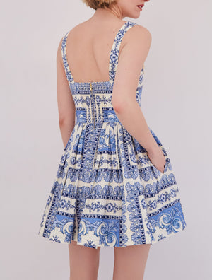 A woman stands facing away, wearing the Cara Cara Micaela Mini Dress—a blue and white patterned fit-and-flare style with a structured bodice, square back, and visible zipper.