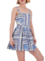 A woman wears the Cara Cara Micaela Mini Dress, featuring a fit-and-flare silhouette, sleeveless square neck, blue and white ornate print, structured bodice, flared skirt, and side seam pockets.