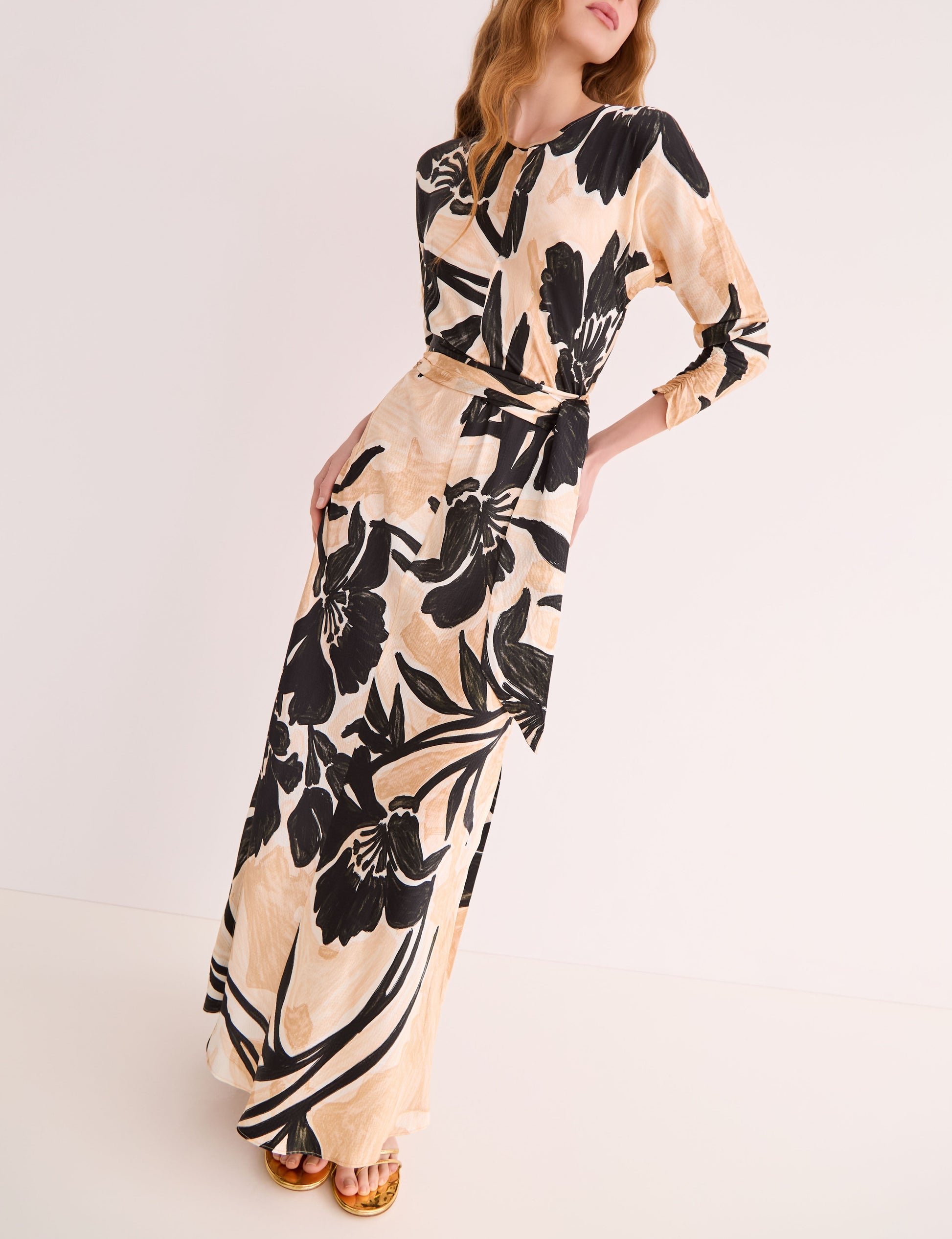 A woman wears the Cara Cara Montgomery Silk Midi Dress, a long-sleeved hammered silk piece featuring an allover beige and black floral print, standing against a plain light background.