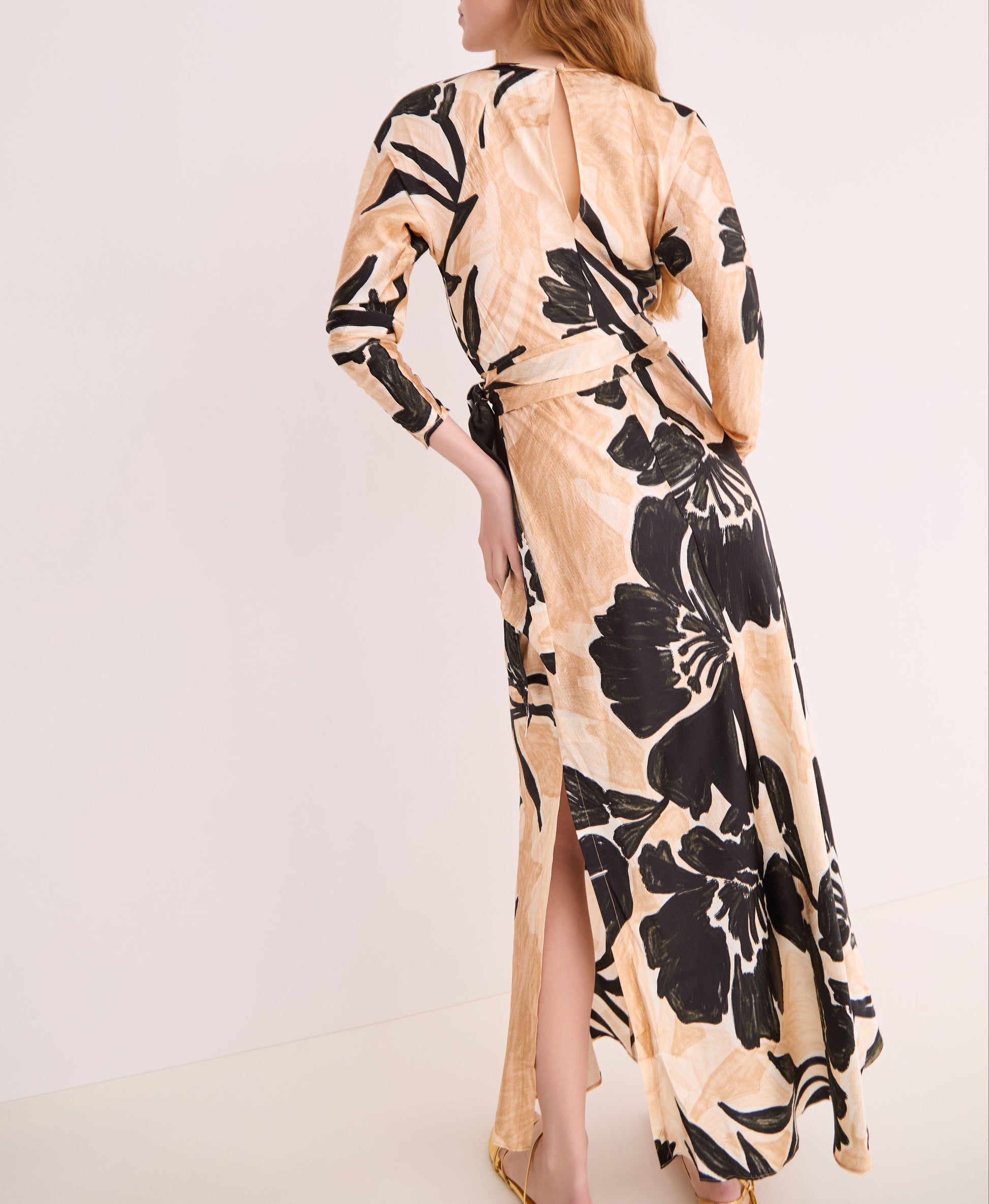 A woman models the Cara Cara Montgomery Silk Midi Dress, featuring hammered beige silk with an allover black floral print and a slit, set against a plain light background.