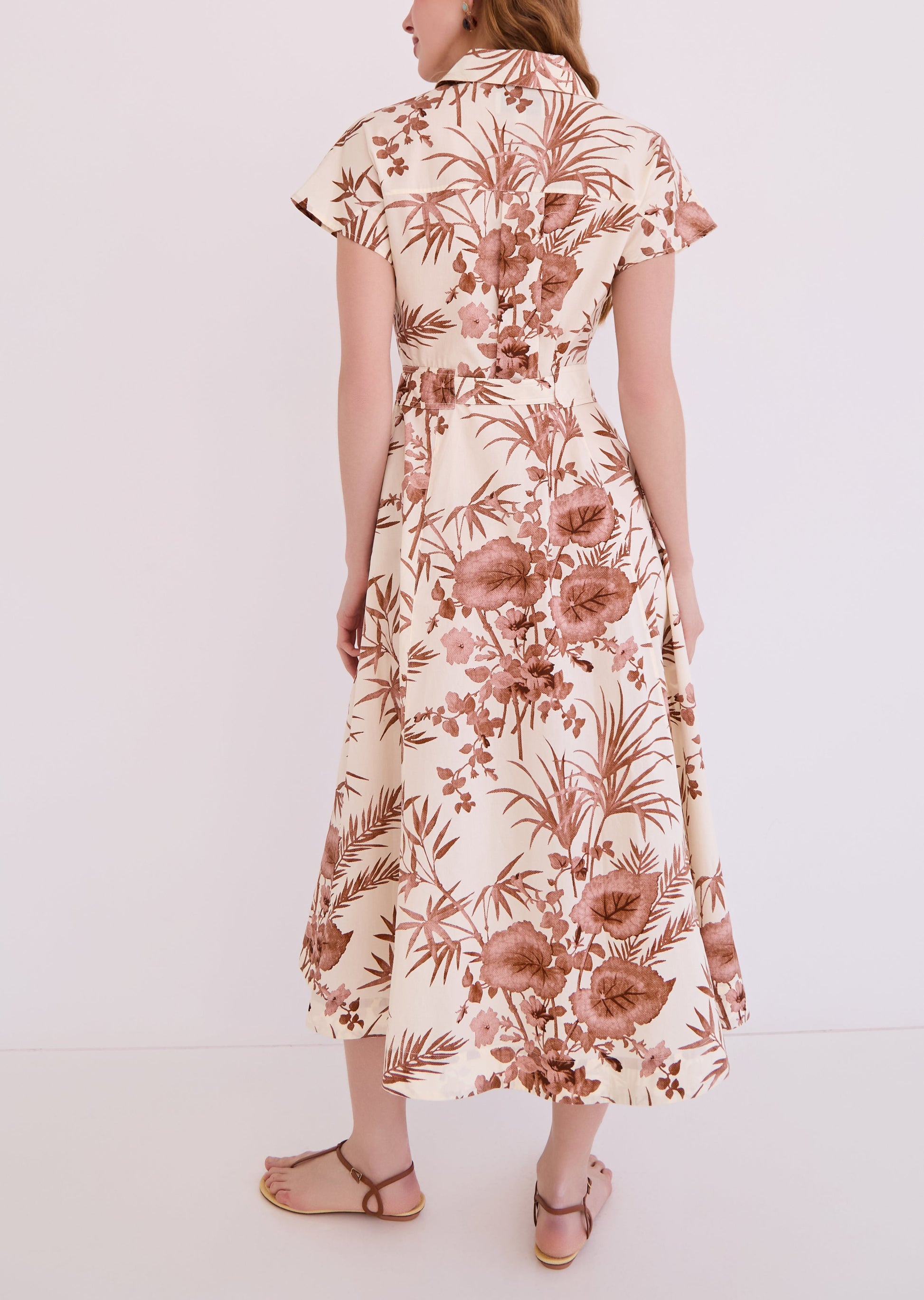A woman faces away, wearing the Cara Cara Sawyer Dress—a cream belted A-line with short sleeves and a brown floral and leaf print—paired with flat sandals.