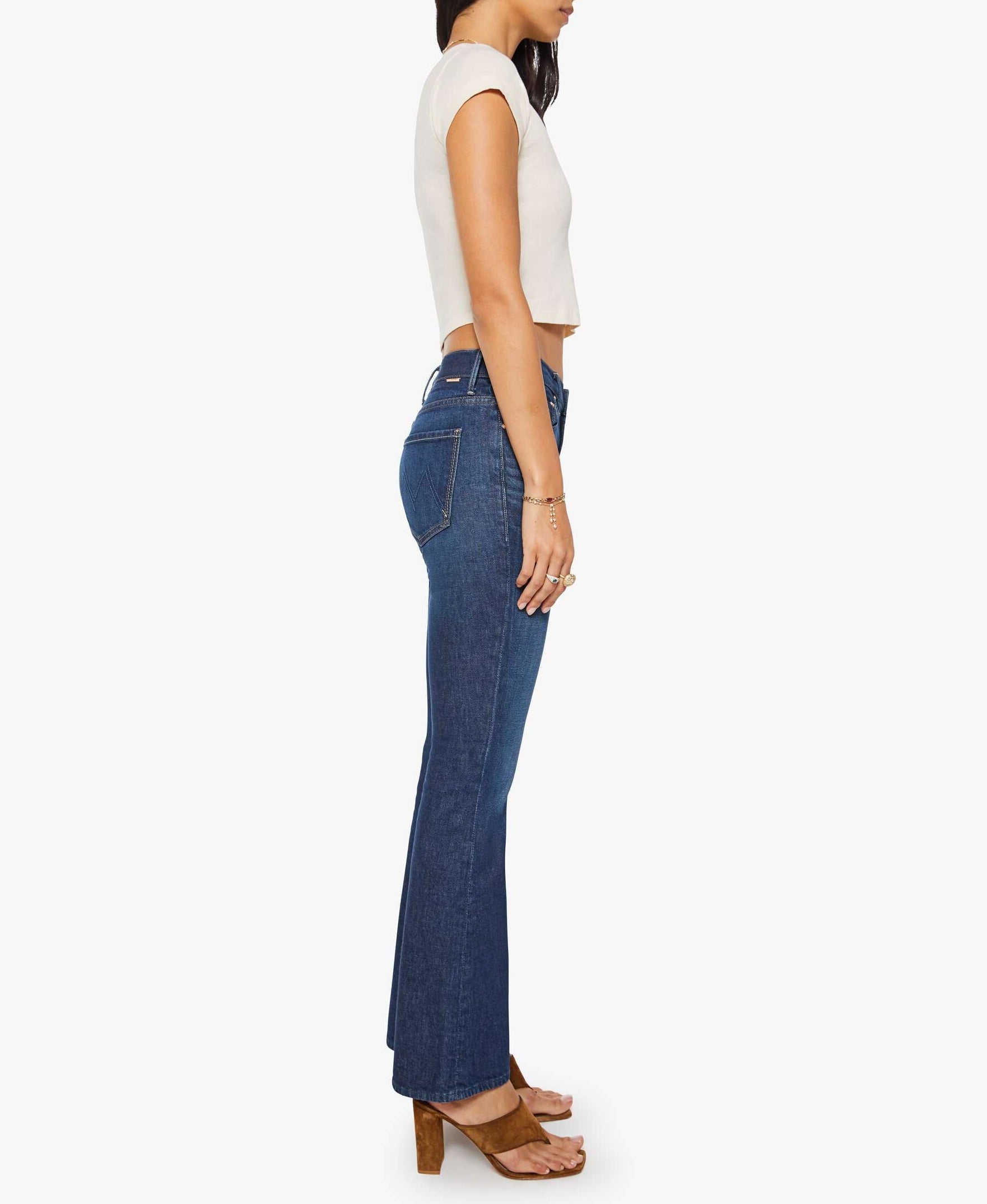 A woman stands in profile against a plain white background, wearing The Weekender mid-rise high-waisted flare jeans by Mother, paired with a white short-sleeve crop top and brown heeled sandals.