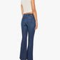 A woman wearing a short-sleeve cream top, brown heeled sandals, and The Weekender mid rise blue flare jeans by Mother in stretch denim with back pockets stands facing away from the camera.