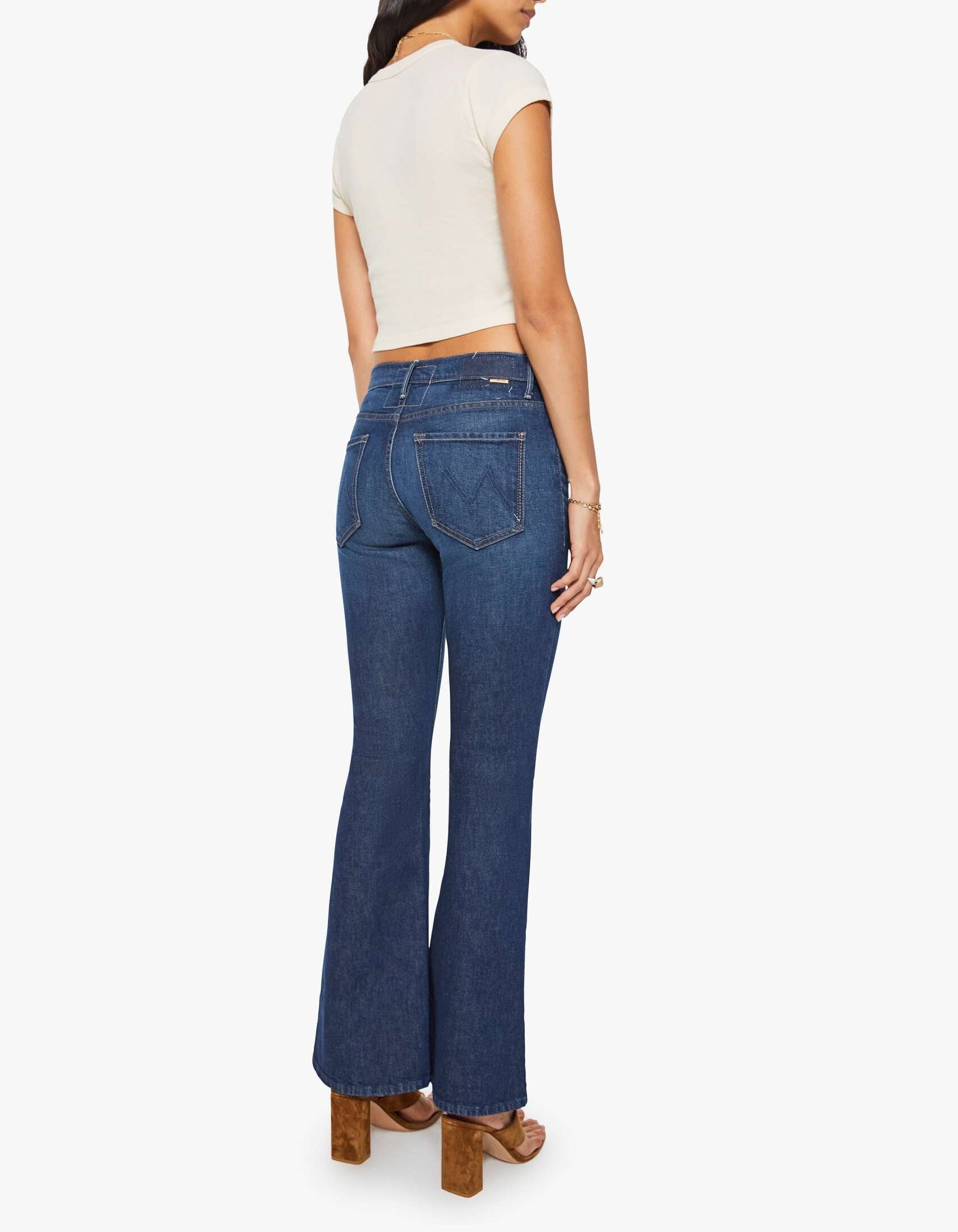 A woman wearing a short-sleeve cream top, brown heeled sandals, and The Weekender mid rise blue flare jeans by Mother in stretch denim with back pockets stands facing away from the camera.
