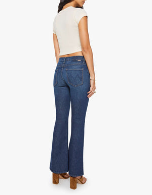 A woman wearing a short-sleeve cream top, brown heeled sandals, and The Weekender mid rise blue flare jeans by Mother in stretch denim with back pockets stands facing away from the camera.