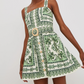 A woman wears the Cara Cara Candy Mini Dress, a sleeveless fit-and-flare mini with green ornate patterns, gold buttons, a square neckline, and a decorative belt buckle.