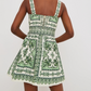 A person shown from the back wearing the Cara Cara Candy Mini Dress, a sleeveless fit-and-flare mini dress featuring green ornate patterns on a white background.