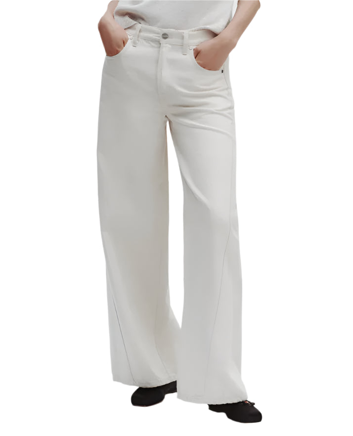 A person stands with hands in pockets against a plain background, wearing the TWP Almost Famous Pant in white, paired with a white top and black shoes.