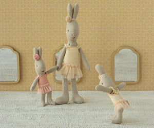 Three Maileg Rabbit, Micro - Ballet Suit & Skirt Cream dolls from Maileg stand on a textured surface, each in a cream ballet outfit with tulle skirts. Patterned wallpaper and small mirrors add charm to the scene.
