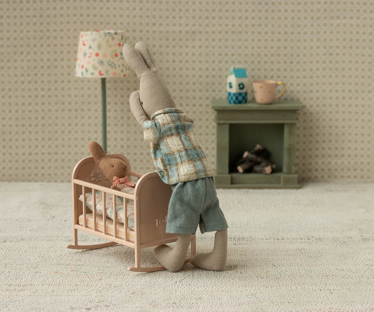 Two Maileg Rabbit, Size 1 - Classic, wearing shirt & shorts in soft fabrics, are in a tiny room; one stands by the crib while the other lies under a blanket. A small lamp and fireplace add charm to this cozy rabbit scene.