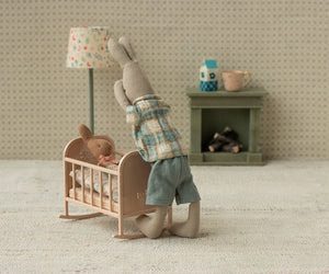 Two Maileg Rabbit, Size 1 - Classic, wearing shirt & shorts in soft fabrics, are in a tiny room; one stands by the crib while the other lies under a blanket. A small lamp and fireplace add charm to this cozy rabbit scene.