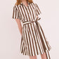 A woman wears the CARA Lagos Dress by Cara Cara, featuring short sleeves, vertical brown and white stripes, a collared neckline, and a matching waist tie, standing against a plain light background.