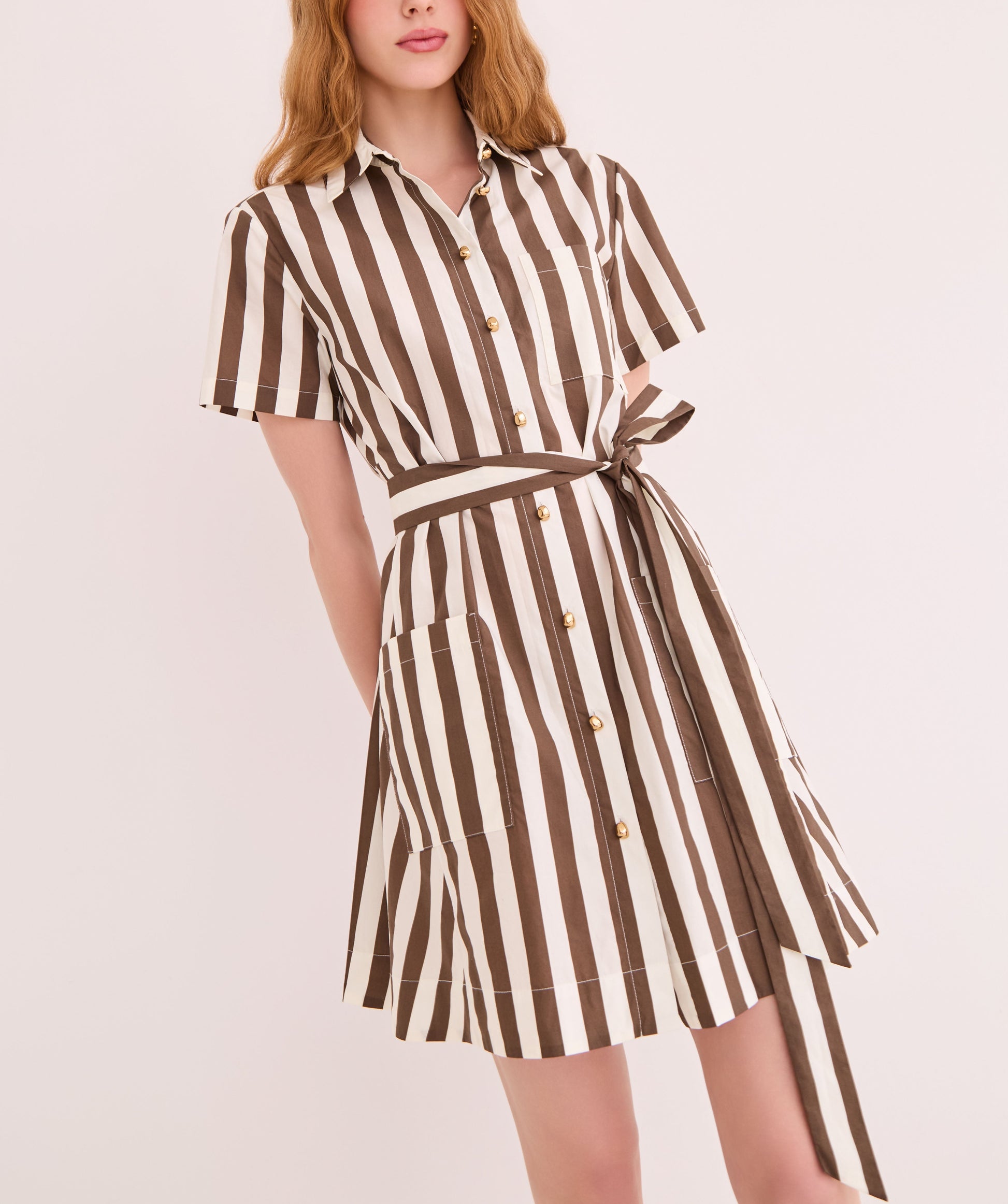 A woman wears the CARA Lagos Dress by Cara Cara, featuring short sleeves, vertical brown and white stripes, a collared neckline, and a matching waist tie, standing against a plain light background.
