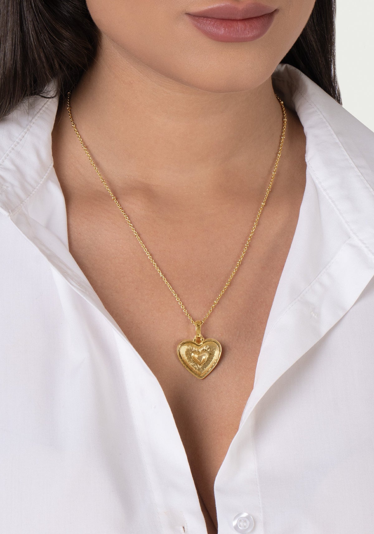 An 18K gold plated brass Heart Radiant Charm hangs from a delicate gold chain worn around a person’s neck.
