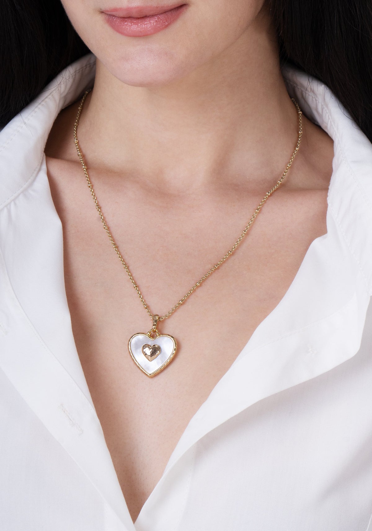 A person wears a gold chain necklace featuring an Anabel Aram Heart Radiant Charm with a white inlay and a smaller central gold heart.