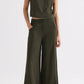 A person stands against a plain background wearing the TWP High Neck Hidden Placket Vest in olive green coated viscose linen with wide-leg trousers, one hand in their pocket and a neutral expression.