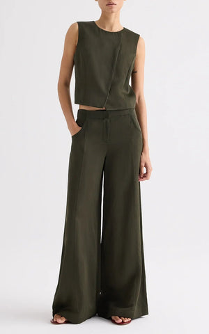 A person stands against a plain background wearing the TWP High Neck Hidden Placket Vest in olive green coated viscose linen with wide-leg trousers, one hand in their pocket and a neutral expression.