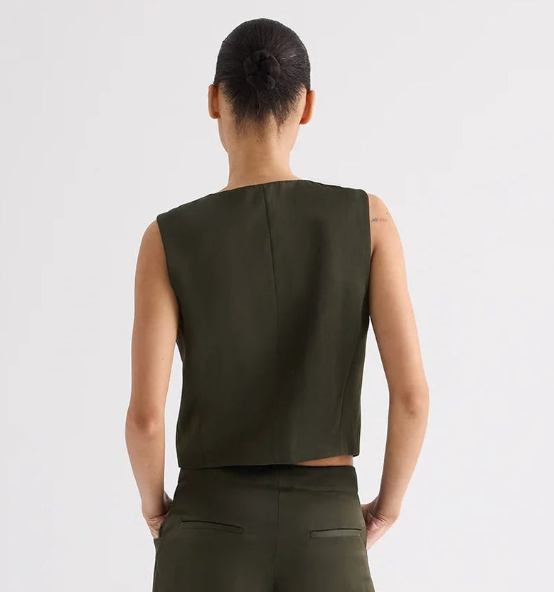 A person stands facing away, wearing the TWP High Neck Hidden Placket Vest in olive green with wide-leg pants that have asymmetric detailing, set against a plain light background.