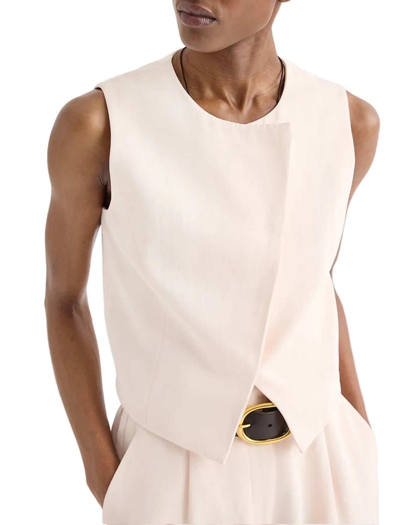 A person wears the TWP High Neck Hidden Placket Vest in light beige with asymmetric detail, paired with matching pleated pants and a black belt featuring a gold buckle.