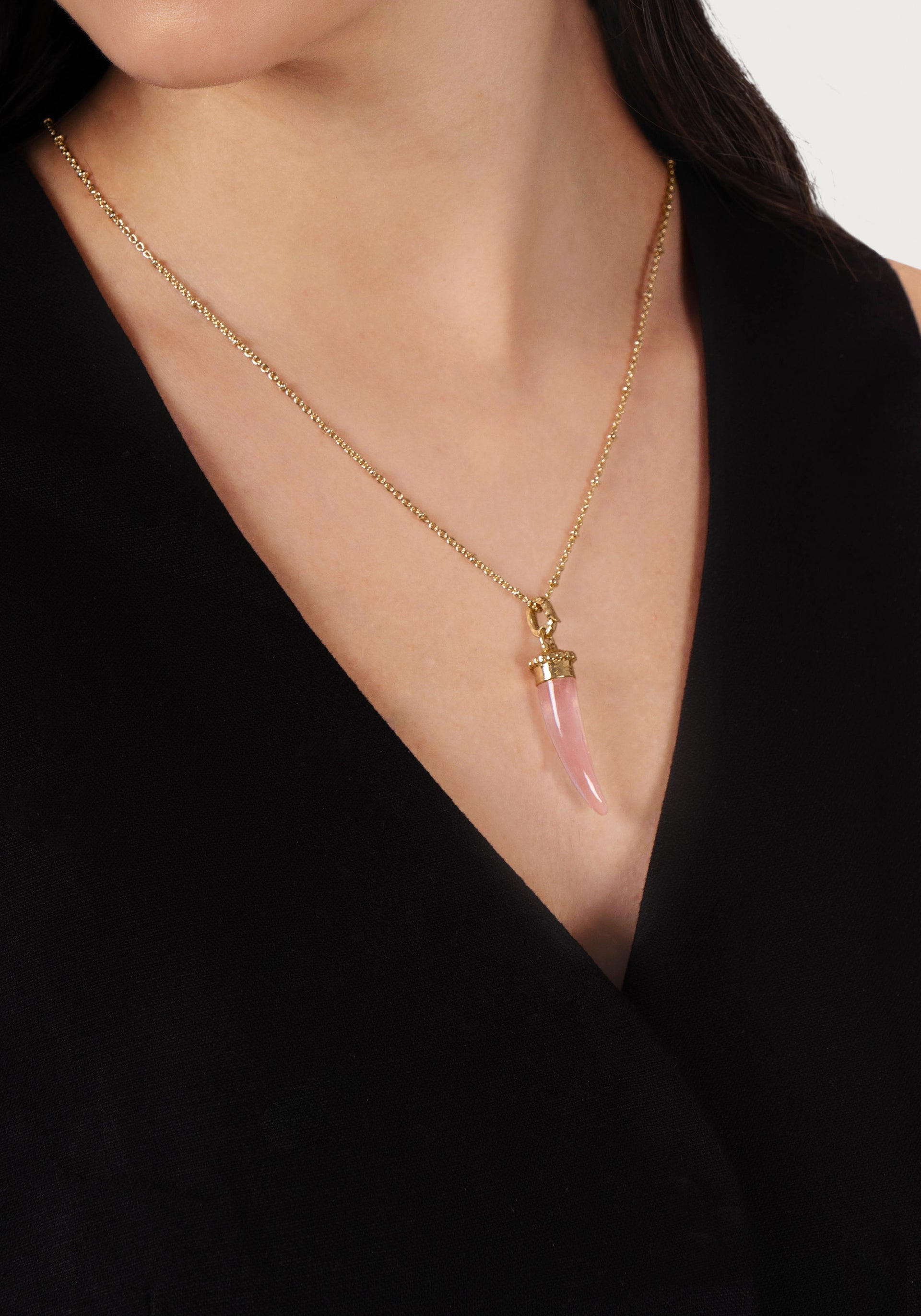 A person wearing the Anabel Aram Milos Stone Horn Charm gold chain necklace with a Mediterranean pink horn pendant, paired with a black V-neck top.