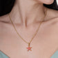 A gold-plated Milos Starfish Charm necklace with a small pink starfish pendant worn around a person’s neck.