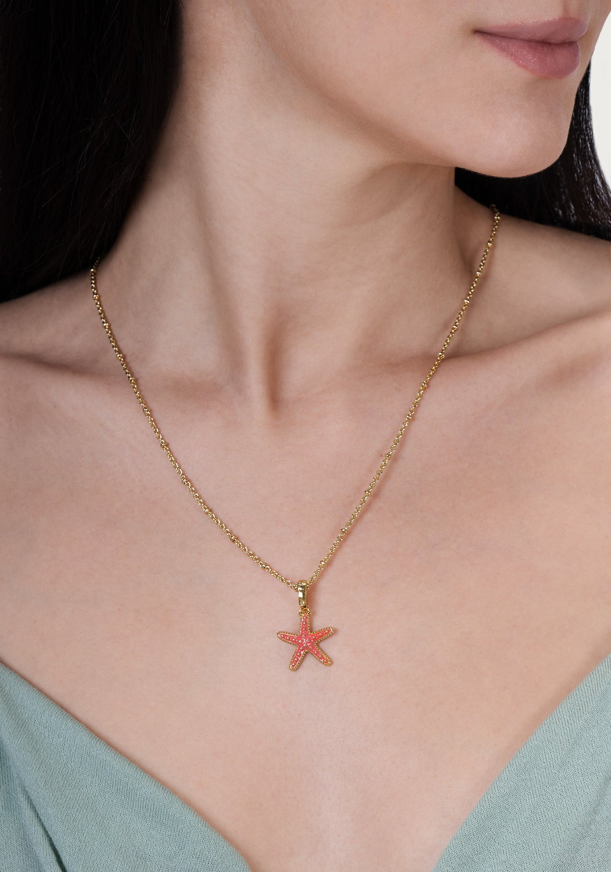 A gold-plated Milos Starfish Charm necklace with a small pink starfish pendant worn around a person’s neck.