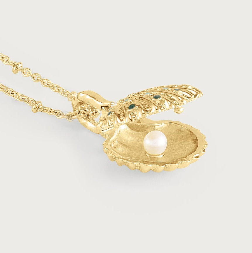 Anabel Aram Milos Shell Locket Charm, a gold shell locket on a chain featuring a single white pearl and small green stones set into the shell exterior.