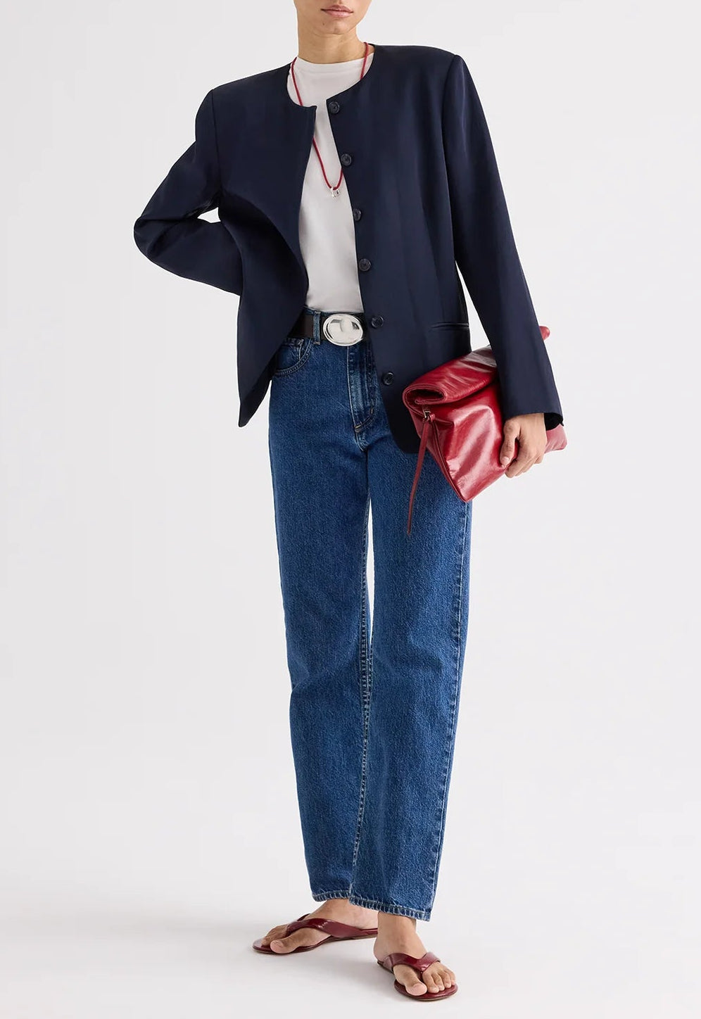A person wearing the TWP Cecelia Coated Viscose Linen Jacket, styled with a white t-shirt, blue jeans, sandals, and holding a large red clutch bag.