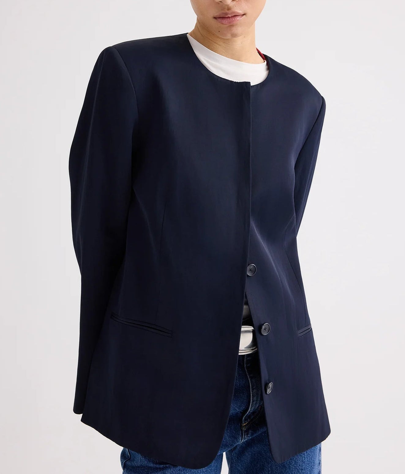 Model wears the TWP Cecelia Coated Viscose Linen Jacket—collarless, navy, with an elongated silhouette—layered over a white t-shirt and blue jeans, set against a plain light background.