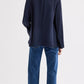 Wearing the TWP Cecelia Coated Viscose Linen Jacket, a navy collarless blazer with an elongated silhouette, blue jeans, and open-toe sandals, the person stands with their back to the camera against a plain light background.