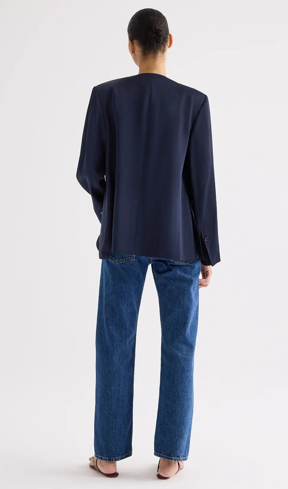 Wearing the TWP Cecelia Coated Viscose Linen Jacket, a navy collarless blazer with an elongated silhouette, blue jeans, and open-toe sandals, the person stands with their back to the camera against a plain light background.
