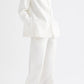 A person stands against a plain white background with hands in their pockets and wearing brown sandals, styled in the TWP Cecelia Linen Jacket—an ivory, collarless blazer with an elongated, loose-fitting silhouette—paired with wide-leg washed linen pants.