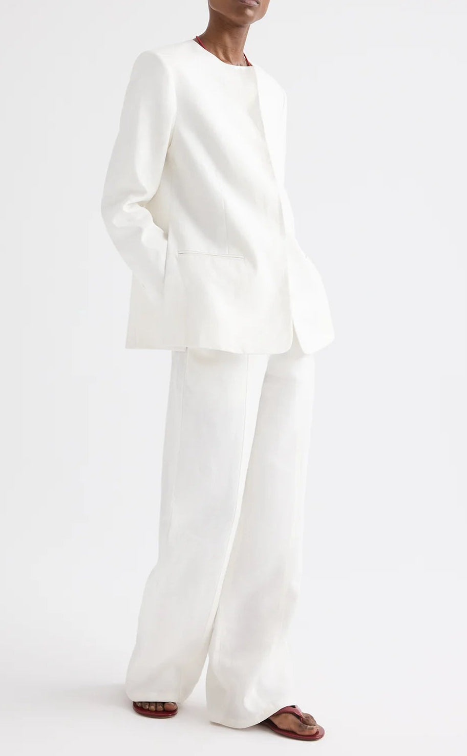 A person stands against a plain white background with hands in their pockets and wearing brown sandals, styled in the TWP Cecelia Linen Jacket—an ivory, collarless blazer with an elongated, loose-fitting silhouette—paired with wide-leg washed linen pants.