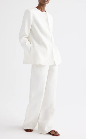A person stands against a plain white background with hands in their pockets and wearing brown sandals, styled in the TWP Cecelia Linen Jacket—an ivory, collarless blazer with an elongated, loose-fitting silhouette—paired with wide-leg washed linen pants.