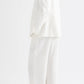 A person wearing the TWP Cecelia Linen Jacket—an ivory washed linen, collarless blazer—paired with wide-leg trousers, stands against a plain white background, facing away from the camera.