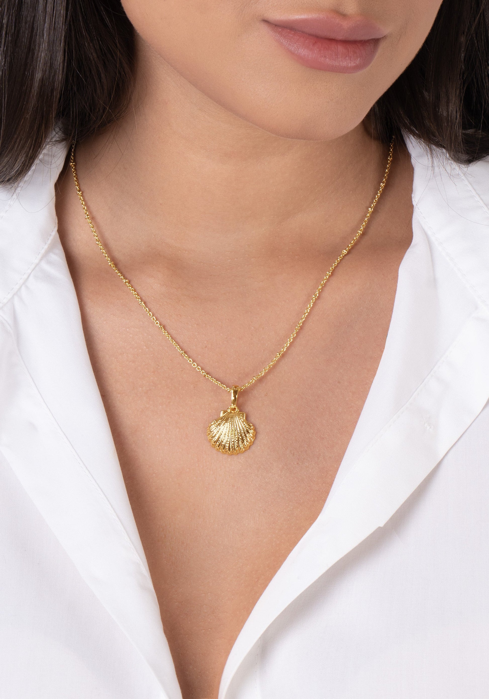 A woman wears a white blouse and the Anabel Aram Scallop Shell Charm, an 18k gold plated necklace.