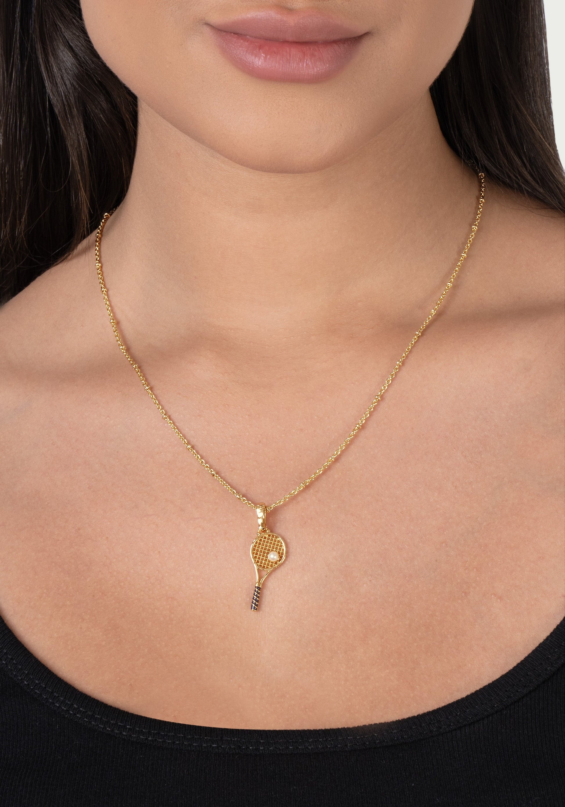 A person wearing the Anabel Aram Tennis Racket Charm gold plated necklace, shown from the shoulders up against a plain background.